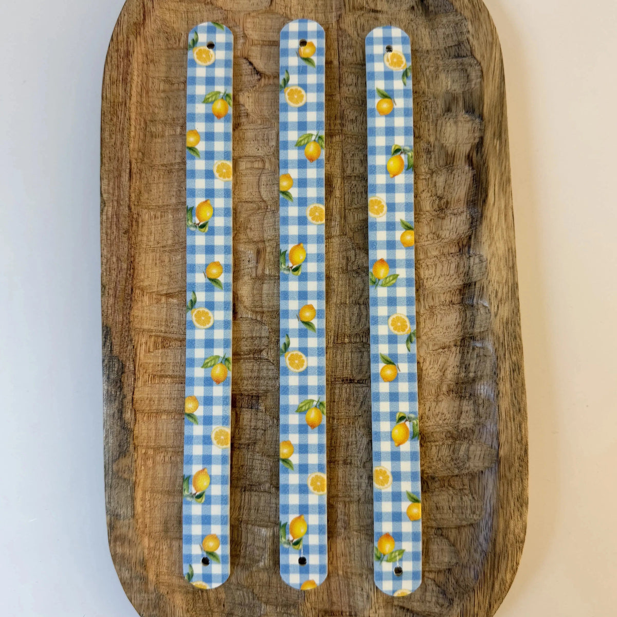 Vue de dessus à plat de trois bandes en biothane pour collier de chien, avec un motif interchangeable chien de carreaux vichy bleu et de citrons jaunes vibrants, sur un plateau en bois.