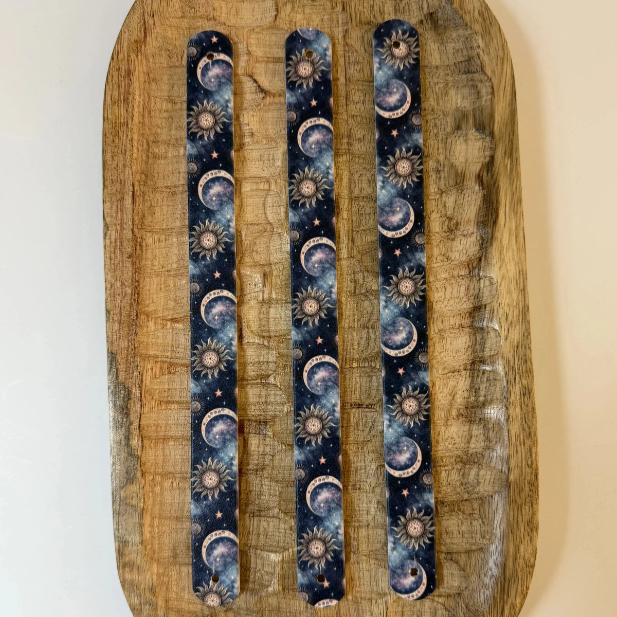Vue de dessus de trois sangles en biothane imprimées pour collier de chien, posées à plat sur un plateau en bois, présentant un motif interchangeable chien cosmique avec lunes, soleils et constellations célestes.