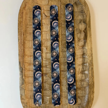 Vue de dessus de trois sangles en biothane imprimées pour collier de chien, posées à plat sur un plateau en bois, présentant un motif interchangeable chien cosmique avec lunes, soleils et constellations célestes.