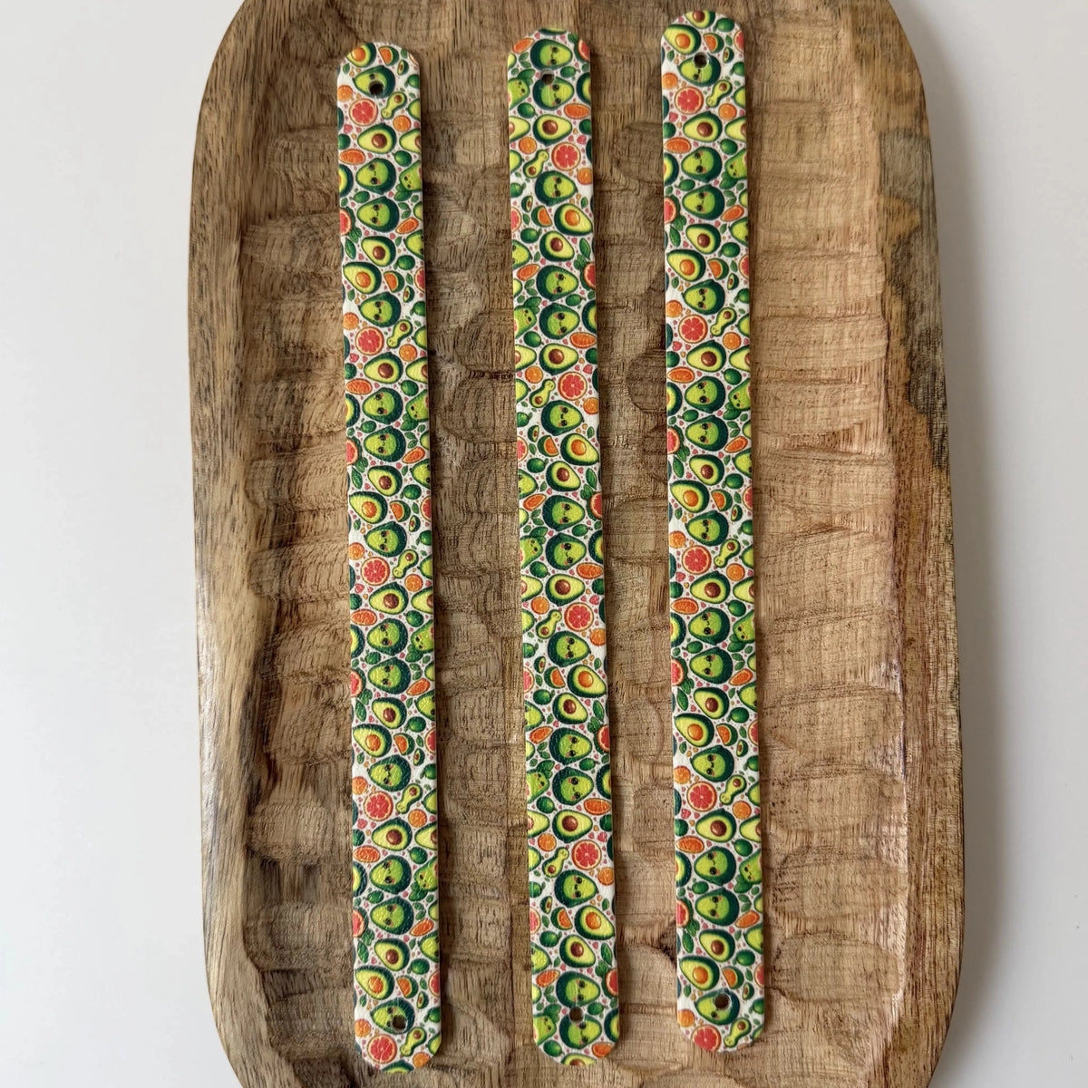 Vue de dessus de trois bandes à motifs interchangeables pour collier de chien en sangle étanche, motif petits avocats rigolos, présentées sur un plateau en bois sculpté.