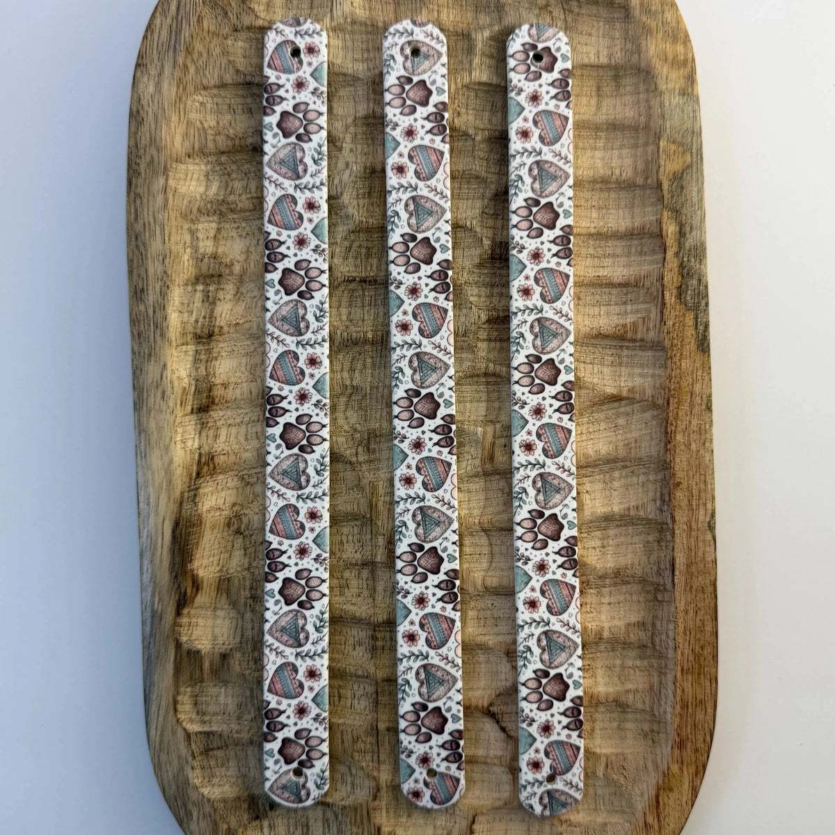 Vue de dessus de trois bandes à motifs interchangeables pour collier de chien en sangle étanche, motif pattes de chien et cœurs style bohème, présentées sur un plateau en bois sculpté.