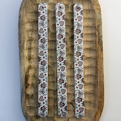 Vue de dessus de trois bandes à motifs interchangeables pour collier de chien en sangle étanche, motif pattes de chien et cœurs style bohème, présentées sur un plateau en bois sculpté.