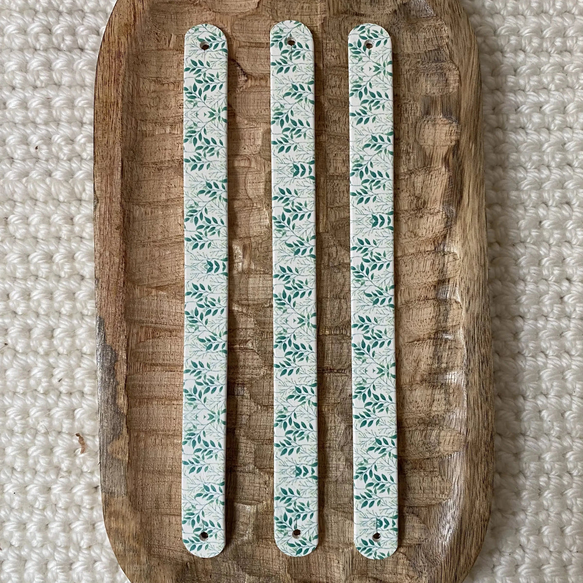 Vue de dessus à plat de trois bandes en biothane pour collier de chien, illustrées d'un motif interchangeable chien botanique de fines feuilles vertes sur fond blanc pur, posées sur du bois.