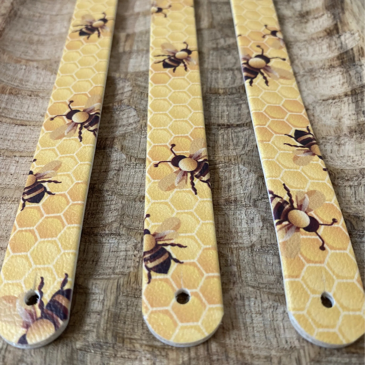 Perspective rapprochée sur trois bandes en biothane posées sur du bois sculpté, décorées d'un motif interchangeable chien chaleureux illustrant des abeilles et un motif d'alvéoles de miel jaune doré.