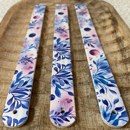 Gros plan en perspective de trois sangles en biothane posées sur un plateau en bois sculpté. Le motif interchangeable chien style aquarelle associe de denses feuillages bleu indigo à des nuances douces de violet et de lavande.