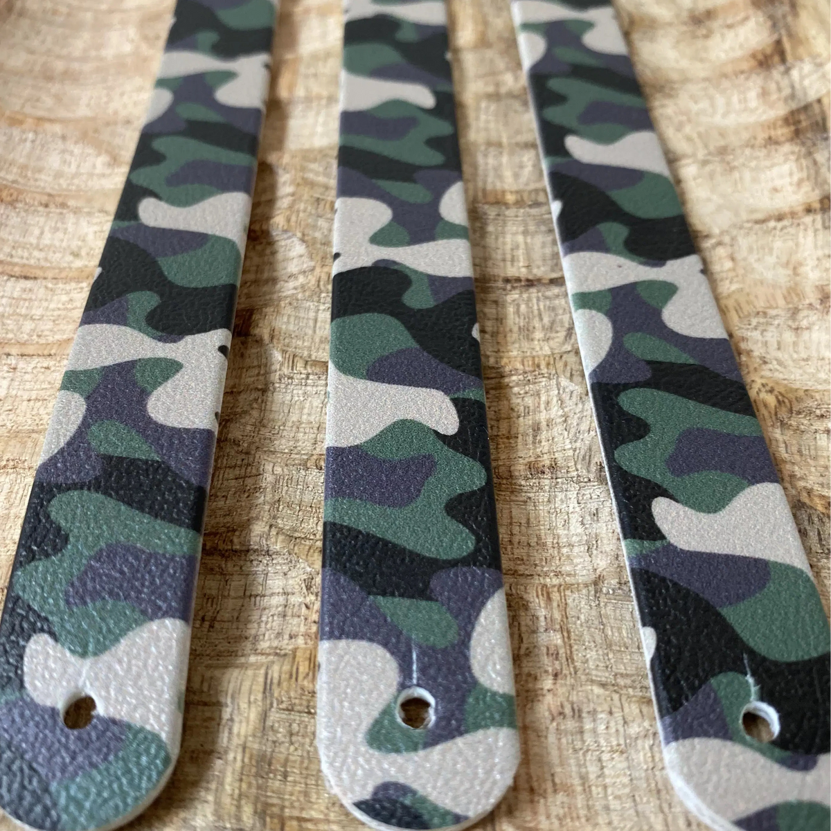 Perspective rapprochée de trois sangles en biothane sur plateau en bois, arborant un motif interchangeable chien camouflage militaire classique aux teintes de vert, kaki, noir et beige.