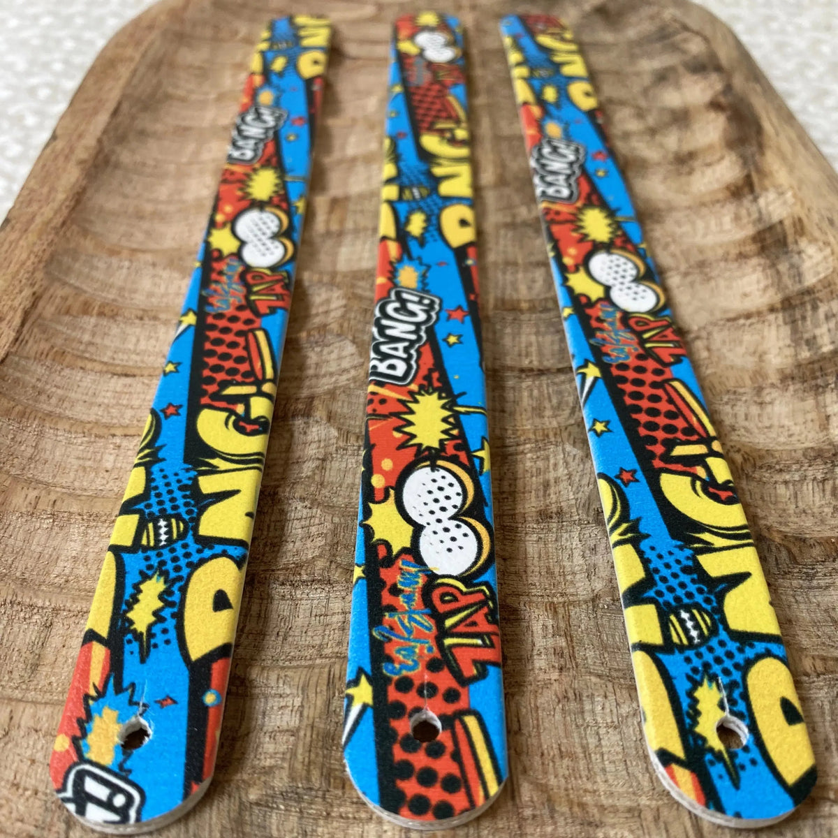 Perspective rapprochée de trois sangles en biothane sur plateau en bois, ornées d'un motif interchangeable chien dynamique style bande dessinée pop art avec les onomatopées BANG! et ZAP! en jaune, rouge et bleu.