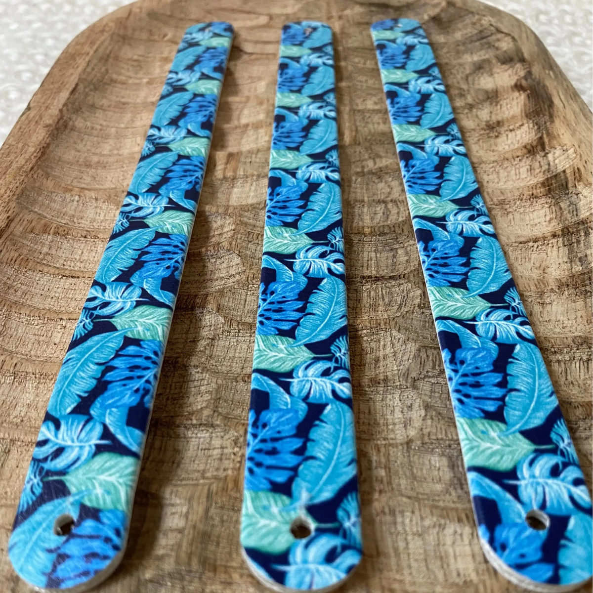 Perspective rapprochée de trois sangles en biothane sur plateau en bois, ornées d'un motif interchangeable chien tropical aux feuilles de monstera et palmes déclinées en bleu cyan et turquoise.