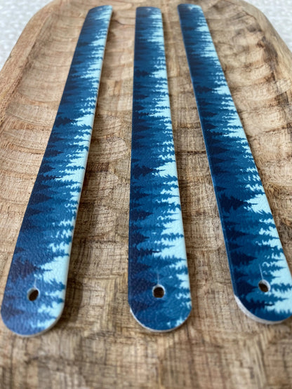 Perspective rapprochée sur trois sangles en biothane posées sur un plateau en bois sculpté, illustrées d'un motif interchangeable chien représentant une forêt de sapins bleus et dégradés d'azur.