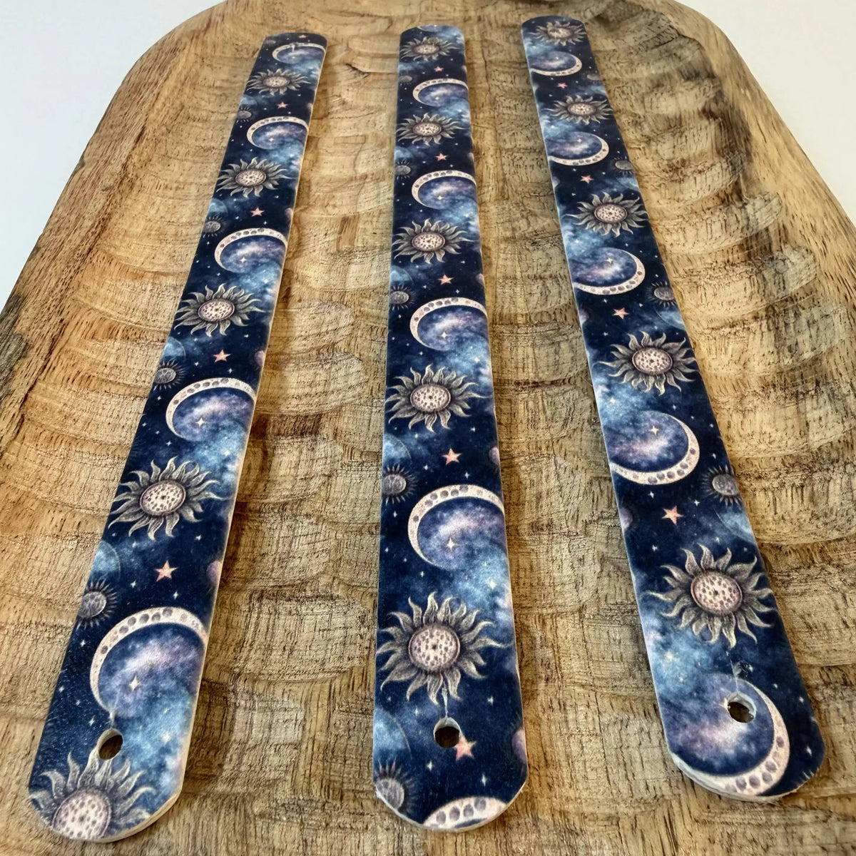 Perspective sur trois bandes de sangle en biothane à motif interchangeable chien sur un plateau en bois sculpté. Elles montrent un design de galaxie cosmique sombre avec des lunes croissantes célestes, des soleils ésotériques et des étoiles sur fond bleu nuit et violet.