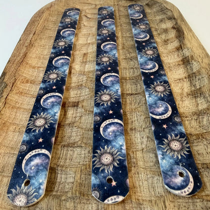 Perspective sur trois bandes de sangle en biothane à motif interchangeable chien sur un plateau en bois sculpté. Elles montrent un design de galaxie cosmique sombre avec des lunes croissantes célestes, des soleils ésotériques et des étoiles sur fond bleu nuit et violet.