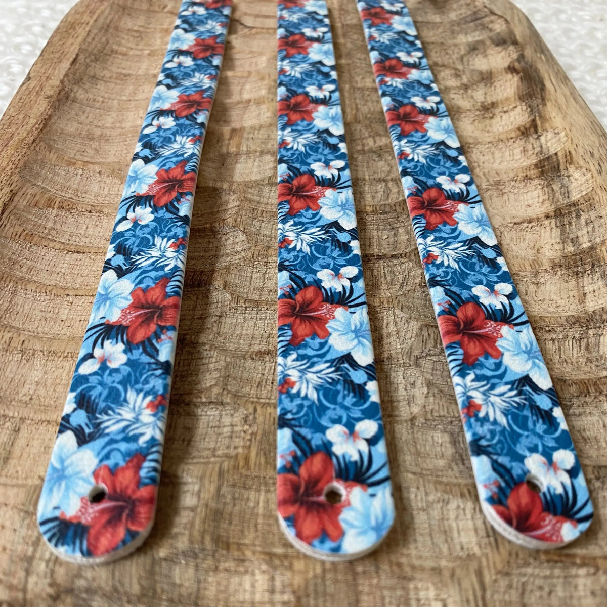 Perspective rapprochée sur trois sangles en biothane posées sur un plat en bois, arborant un motif interchangeable chien exotique aux grandes fleurs d'hibiscus rouges sur fond bleu floral.
