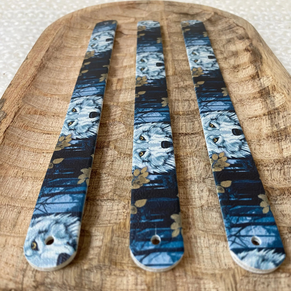Perspective rapprochée sur trois sangles en biothane posées sur un plat en bois, illustrées d'un motif interchangeable chien poétique représentant des têtes de loups au cœur d'une forêt nocturne bleue agrémentée de feuilles dorées.