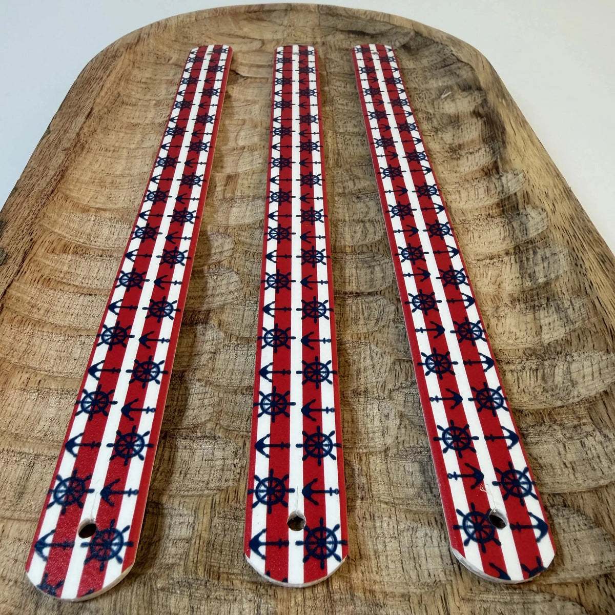Gros plan en perspective de trois bandes en biothane à motif interchangeable chien sur un plateau en bois. Le design présente des rayures horizontales rouges et blanches avec des ancres bleu marine et des barres de gouvernail bleu marine, avec des bords rouges.