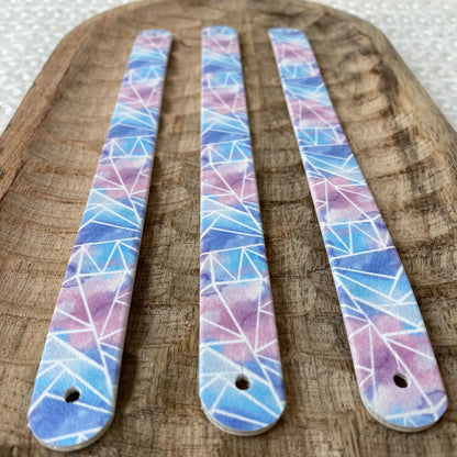 Perspective rapprochée de trois sangles en biothane sur plateau en bois, décorées d'un motif interchangeable chien géométrique effet mosaïque aux nuances d'aquarelle rose, mauve et bleue.
