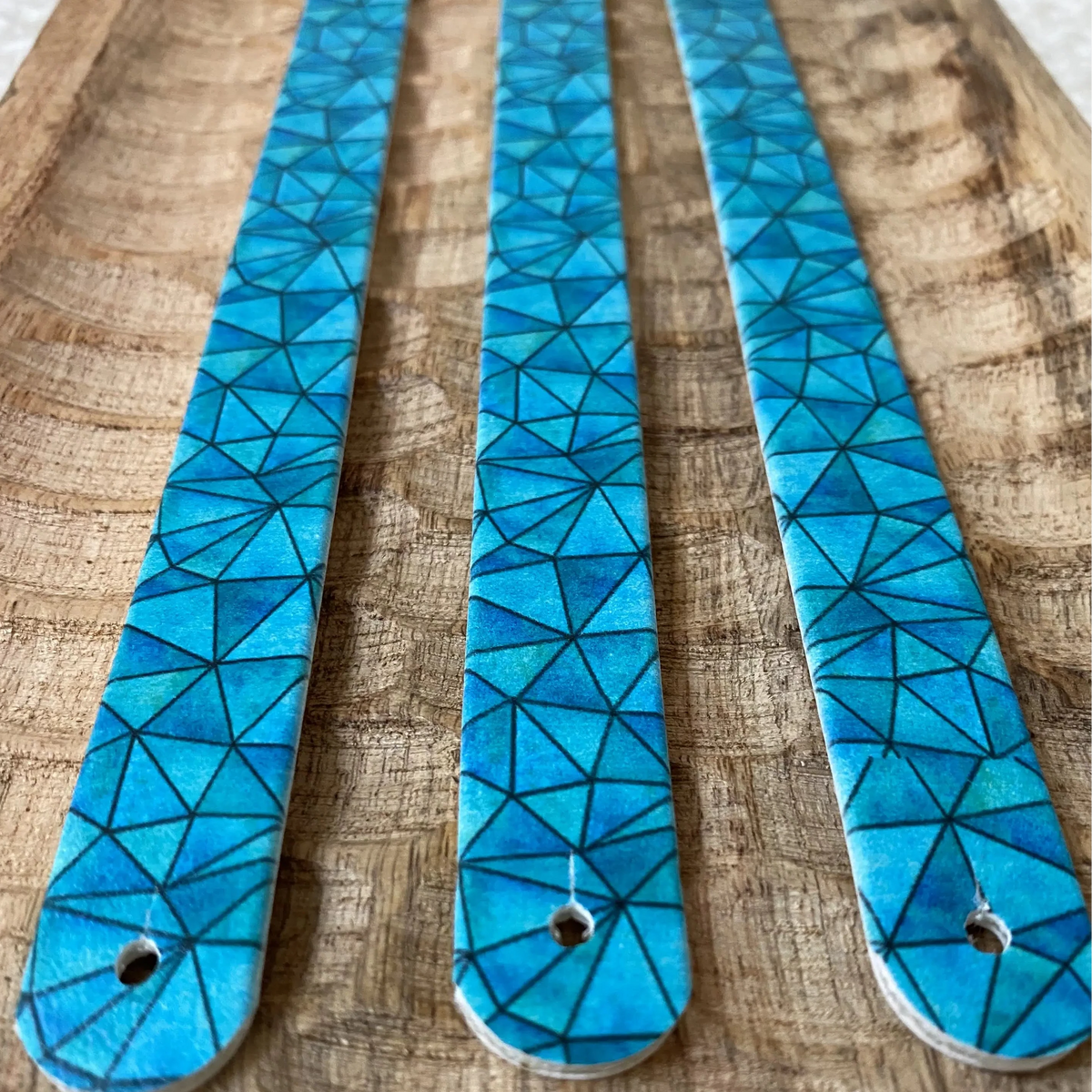 Perspective sur trois sangles en biothane posées sur du bois texturé. Le visuel arbore un motif interchangeable chien géométrique façon vitrail ou mosaïque aux multiples facettes bleu ciel et turquoise.