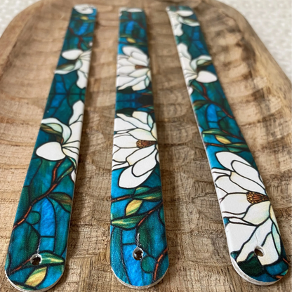 Perspective rapprochée sur trois sangles en biothane posées sur du bois, sublimées par un motif interchangeable chien artistique façon vitrail aux fleurs blanches et nuances bleu-vert.