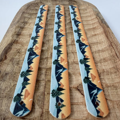 Vue de dessus à plat sur trois sangles en biothane identiques, alignées parfaitement parallèlement sur un plateau en bois. Elles arborent le même motif de paysage de montagne stylisé au coucher du soleil.