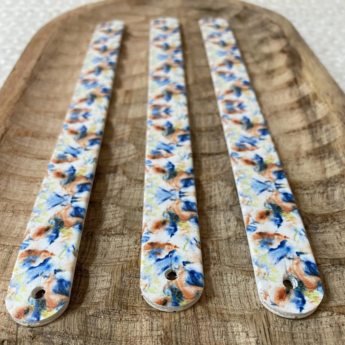 Vue de dessus à plat de trois bandes en biothane pour collier de chien, imprimées d'un motif interchangeable chien de carreaux floraux kaléidoscopiques bleus et oranges, sur un plateau en bois.