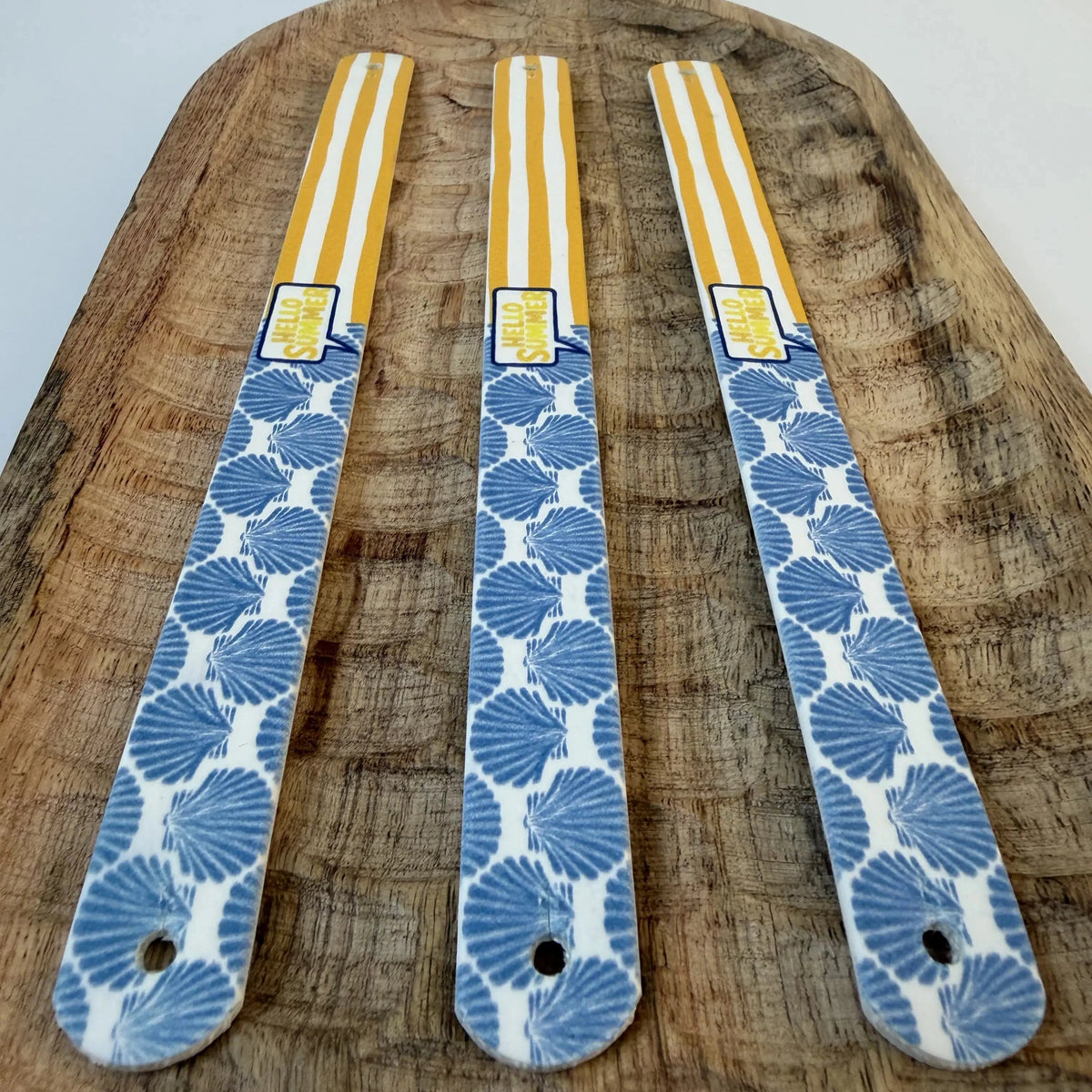 Vue en perspective de trois bandes à motifs interchangeables pour collier de chien en sangle étanche, motif coquillages bleus, rayures jaunes et bulle "Hello Summer", posées sur un plateau en bois sculpté.