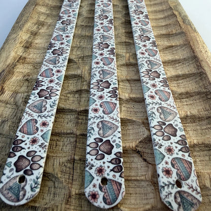 Vue en perspective de trois bandes à motifs interchangeables pour collier de chien en sangle étanche, motif pattes de chien et cœurs style bohème, posées sur un plateau en bois sculpté.
